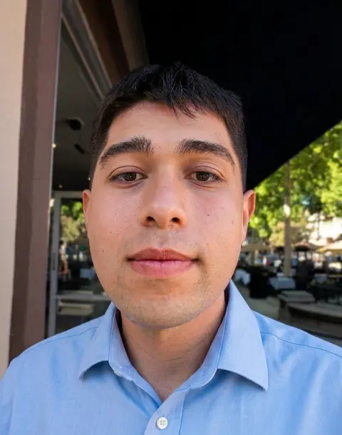 Carlos, smiling, wearing a light blue button-up shirt outdoors.
